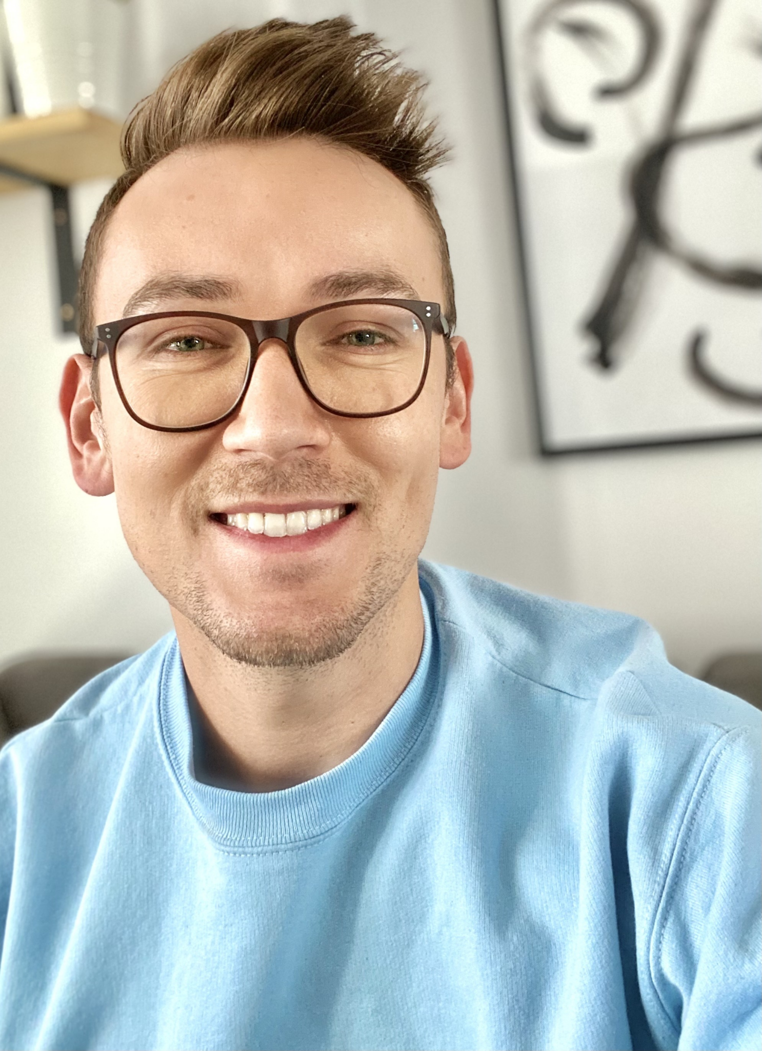 Picture of Kenan Karalic, Web Administrator. A man with light brown hair and brown framed glasses looks at the camera, dressed in a blue sweatshirt. Behind him, framed art and a wooden shelf is visible.