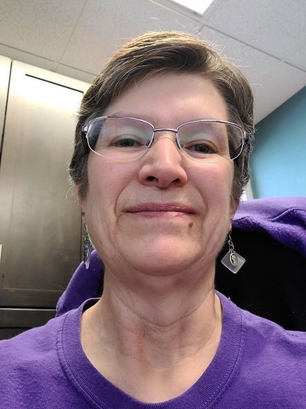 Picture of Melody Gritton, Treasurer. She wears a purple t-shirt and looks slightly downward toward the camera, eyes framed by silver-rimmed glasses.