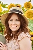 Picture of Randi Beckstrom, Secretary. A woman in a wide-brimmed straw hat and a floral dress stands in a field of sunflowers. She smiles at the camera, reddish brown hair cascading down her shoulders.