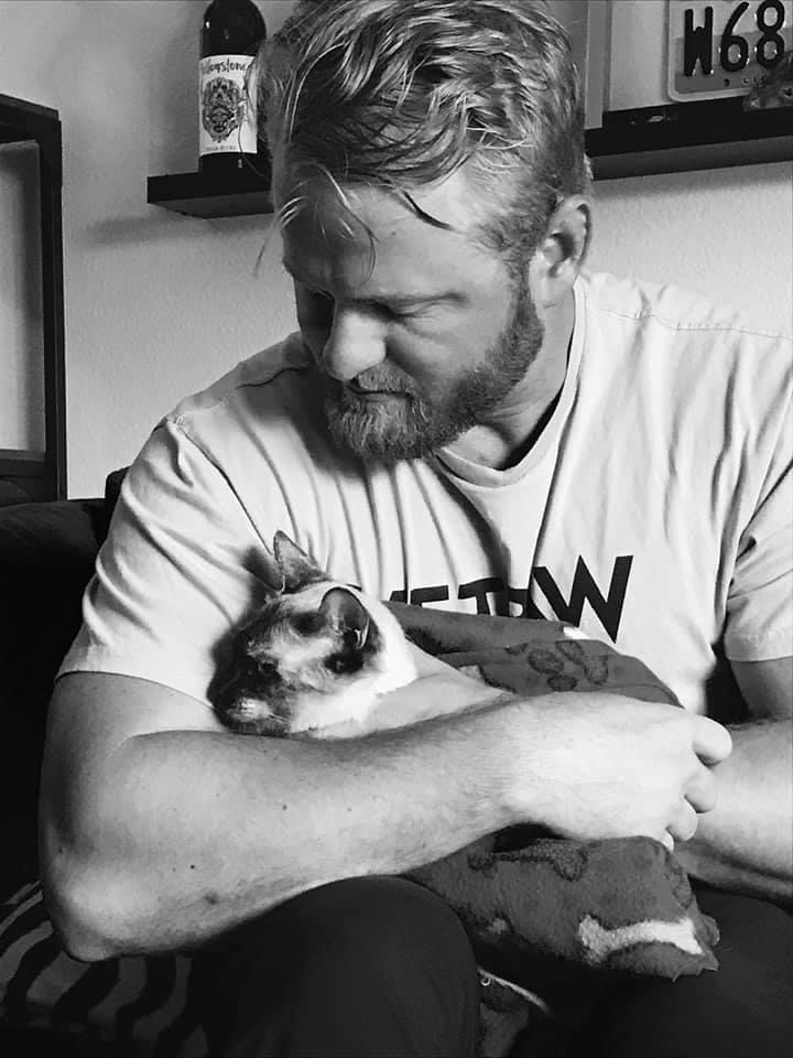 Ryan Tate, looks down at his arms as he cuddles a cat. The cat looks over his elbow, toward the left side of the image.